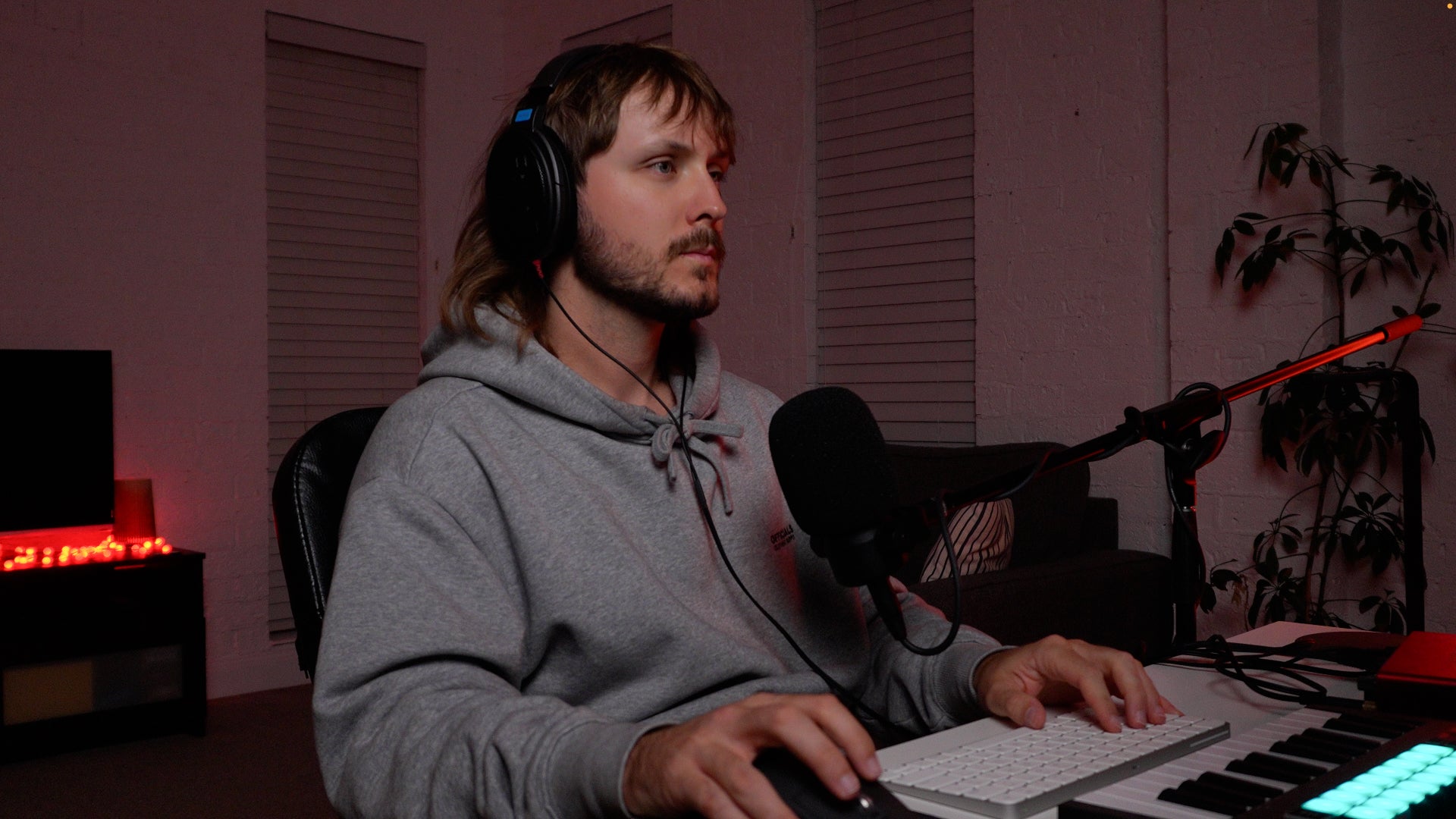 Man in a gray hoodie sitting at a desk with a microphone and computer, in a dimly lit room.