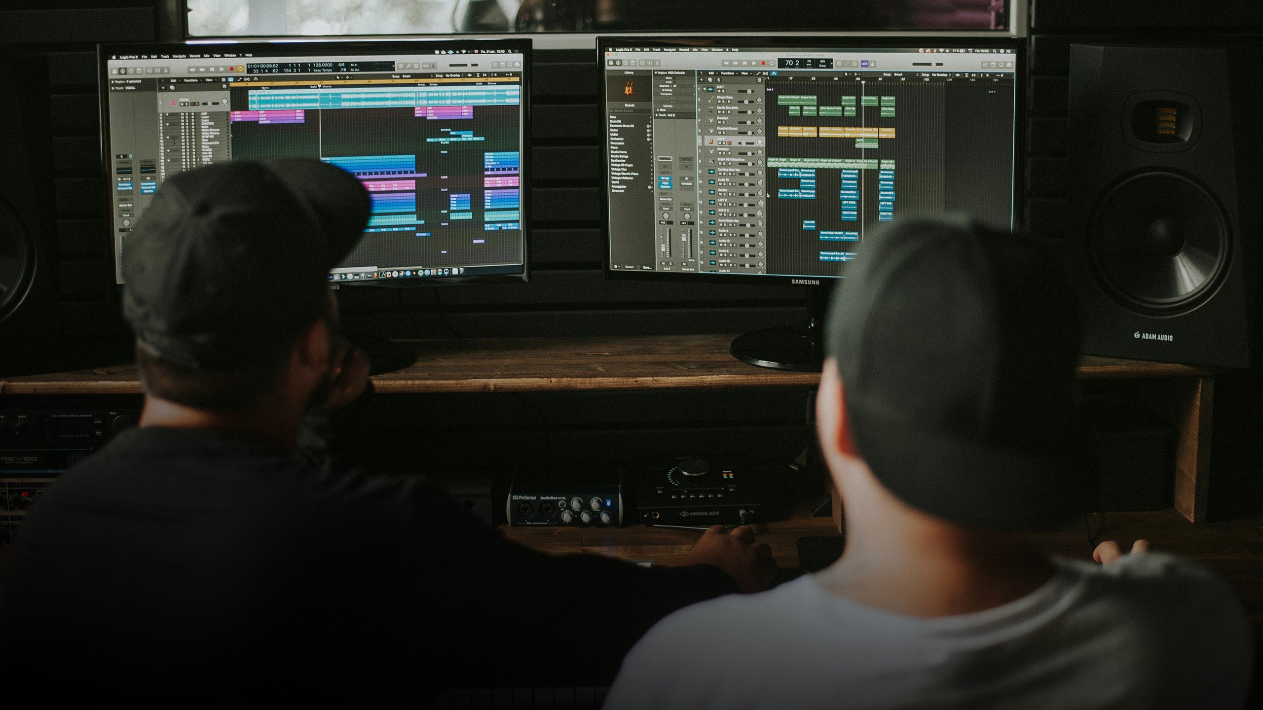 Two people in a recording studio with multiple screens displaying audio software.