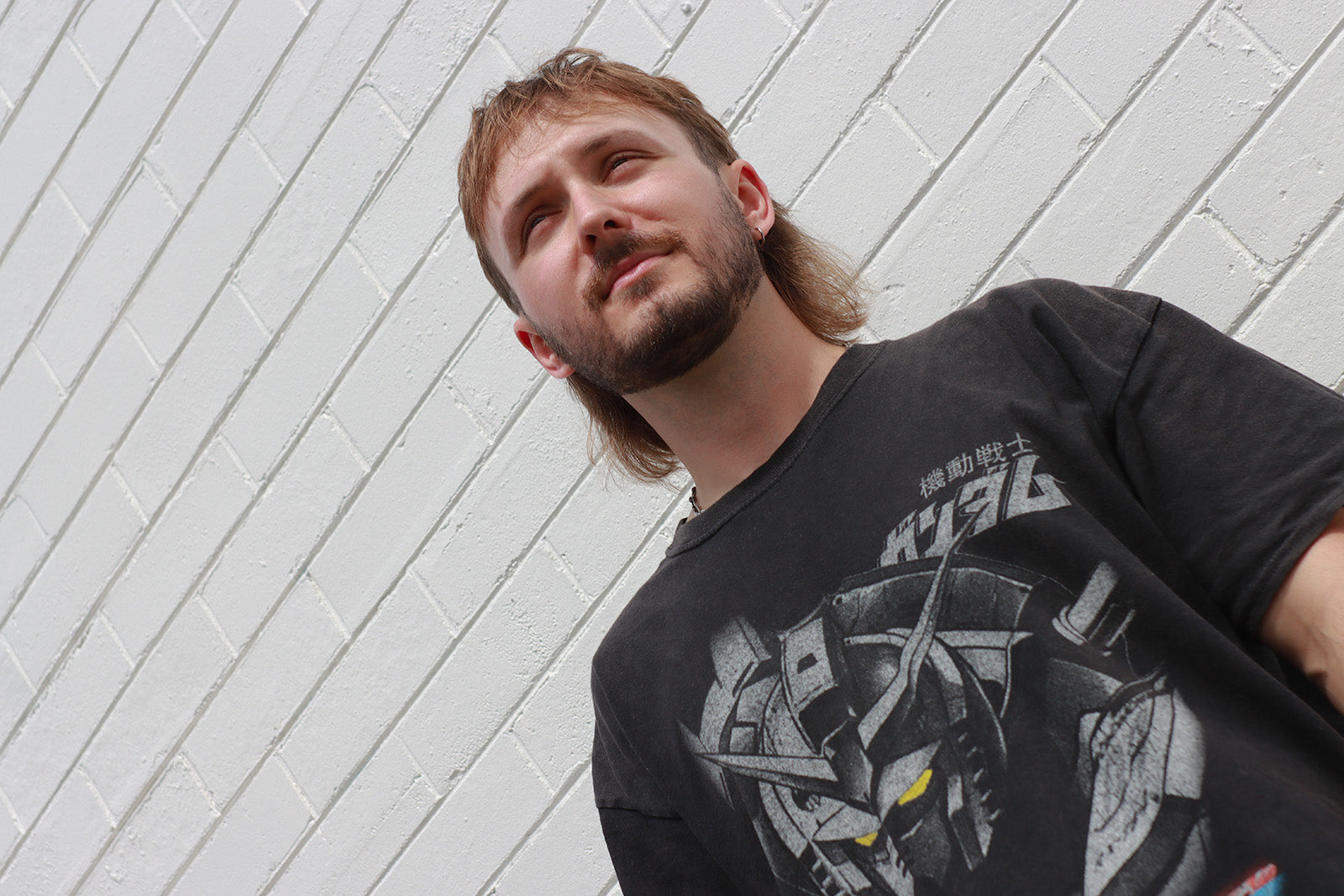 DJ Moiler press shot wearing a black t-shirt with a graphic design against a white brick wall.